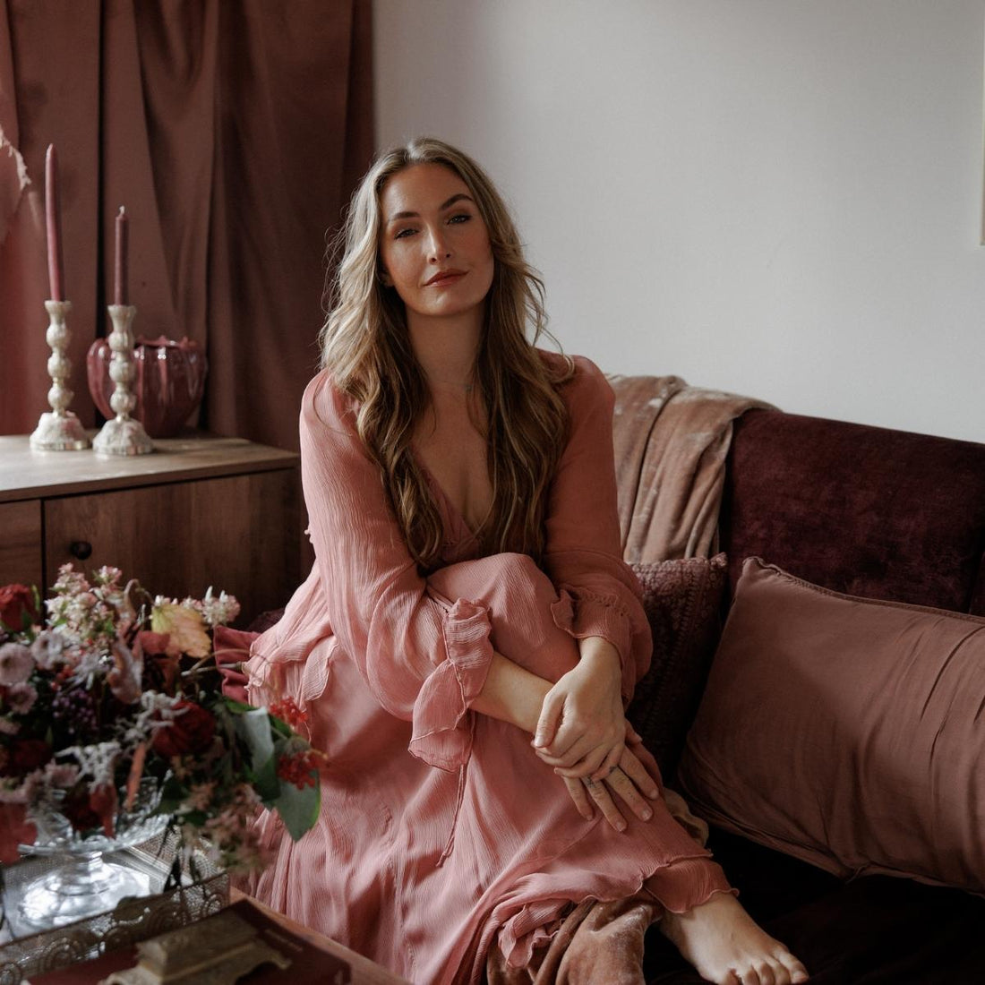 Lara Raybone sits barefoot on a velvet sofa in a soft pink dress, surrounded by warm, earthy tones and flowers, exuding calm, confidence, and grace.