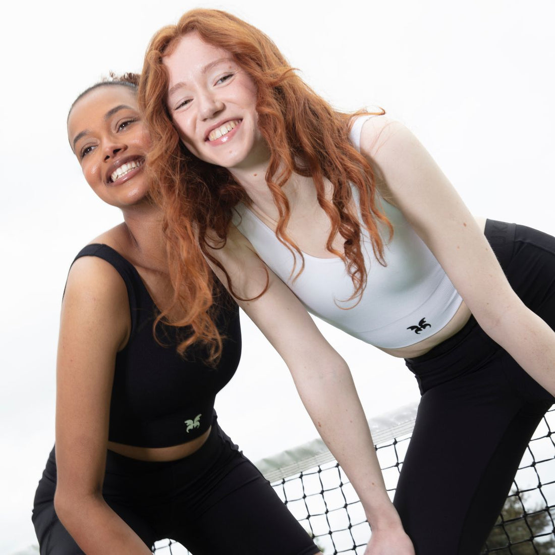 Two smiling teens dressed in activewear, leaning on a tennis net outdoors, representing confidence, friendship, and body positivity during puberty.