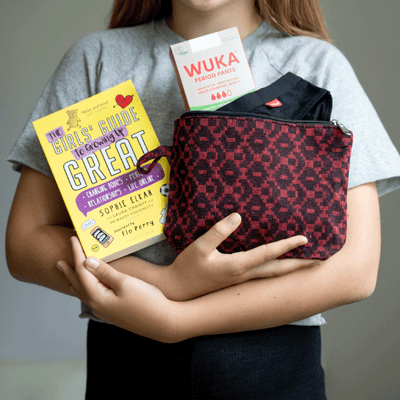 A girl holds her first period pack, it has a book (Girls Guide to growing up great), WUKA period pants, and washbag. 